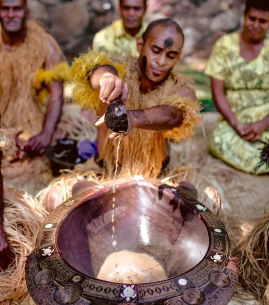 A man in traditional dress pours kava from a coconut shell into a carved communal bowl as others sit together