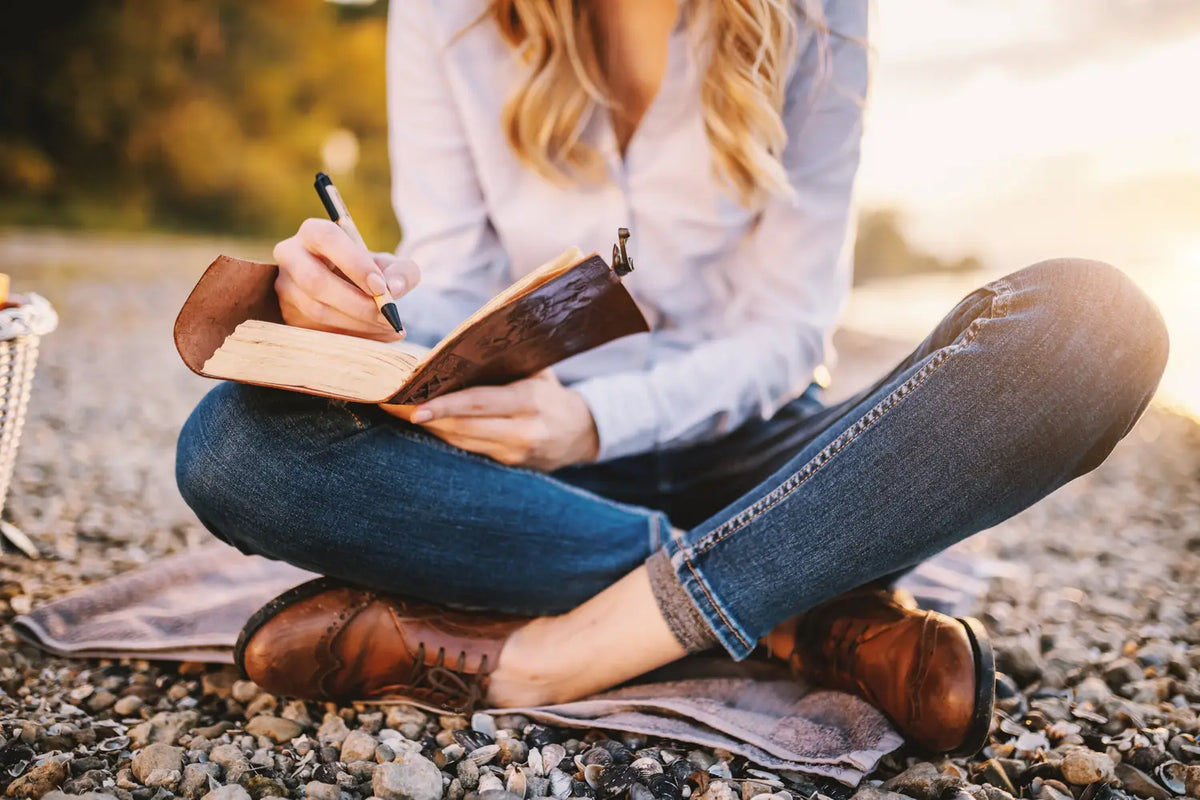 A person writes in a leather journal beside the water at golden hour