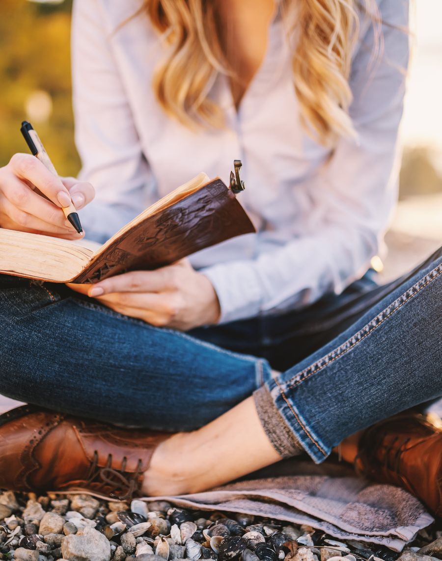 A woman sitting cross-legged on a pebble beach, writing in a leather journal at golden hour